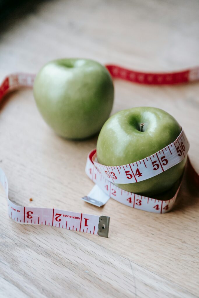Two green apples on a wooden surface wrapped with a red and white measuring tape, symbolizing health and diet.